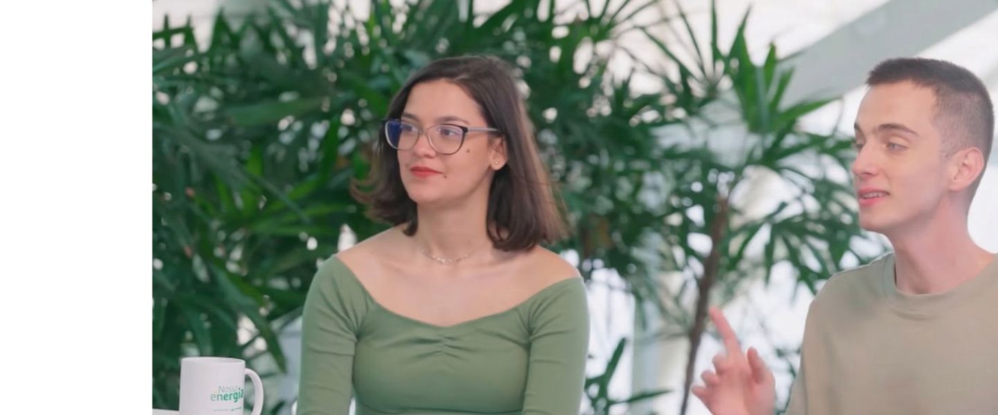 Pessoa com óculos e blusa verde sentada ao lado de outra pessoa que gesticula, em um ambiente interno com plantas ao fundo.