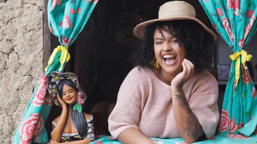 A influenciadora Amora Mota sorri, apoiada em uma janela com cortinas estampadas. Ela usa chapéu de palha e suéter rosa, e ao seu lado há uma escultura de barro de uma mulher.A influenciadora Amora Mota sorri, apoiada em uma janela com cortinas estampadas. Ela usa chapéu de palha e suéter rosa, e ao seu lado há uma escultura de barro de uma mulher.