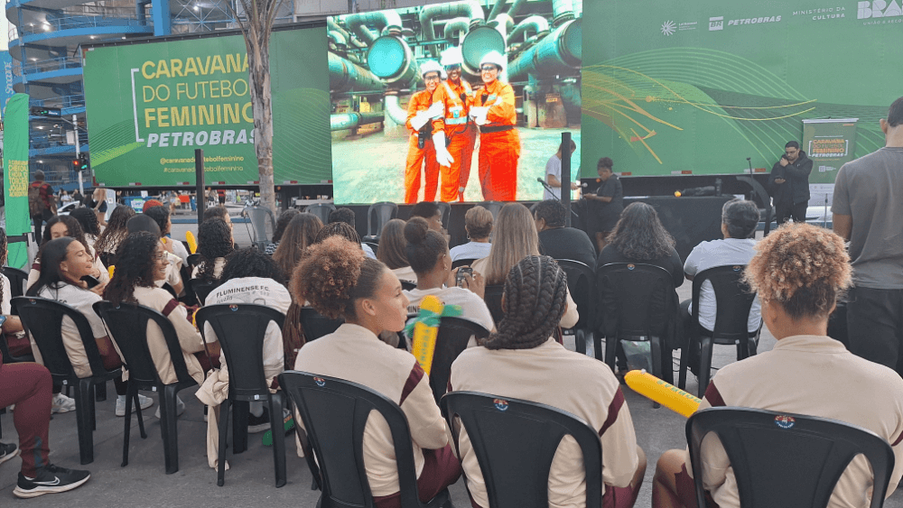 Diversas mulheres jovens sentadas em cadeiras plásticas ao ar livre, em frente a um palco da Caravana de Futebol Feminino Petrobras.