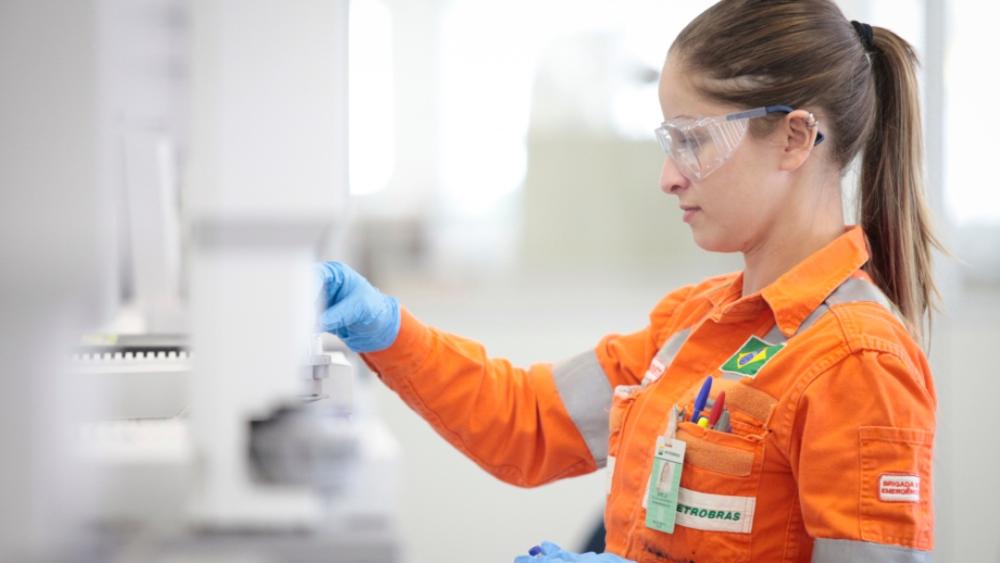 Mulher com uniforme laranja da Petrobras dentro de um laboratório. Ela usa óculos de proteção.