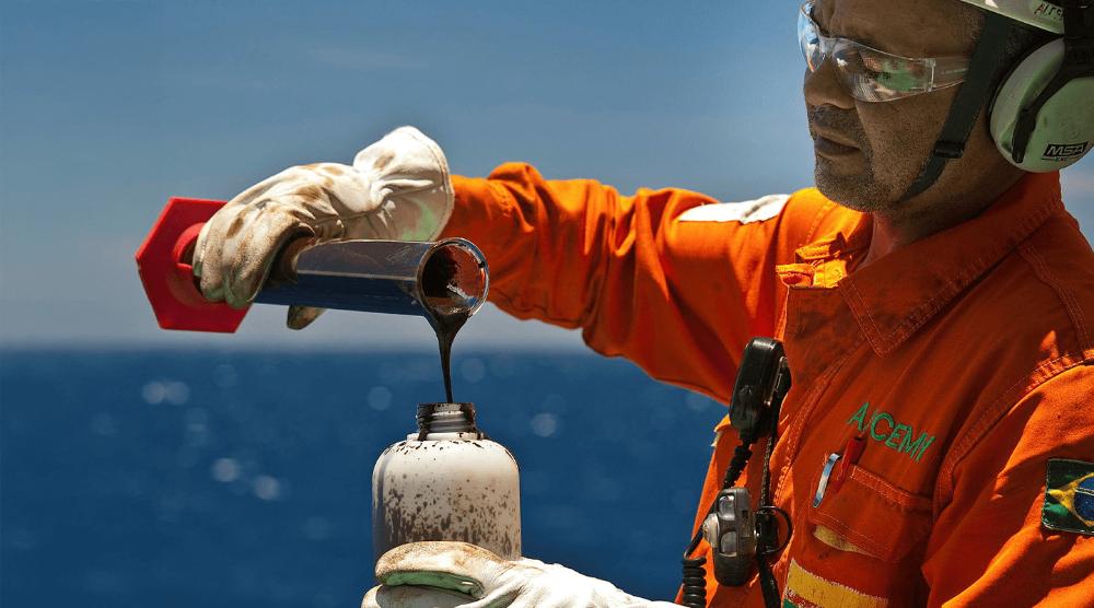 Homem com uniforme da Petrobras e proveta de petróleo com o mar ao fundo.