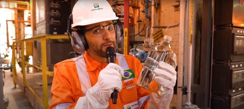 Iberê com uniforme da Petrobras, usando EPI completo, segura um frasco com líquido transparente e fala ao microfone em ambiente industrial.