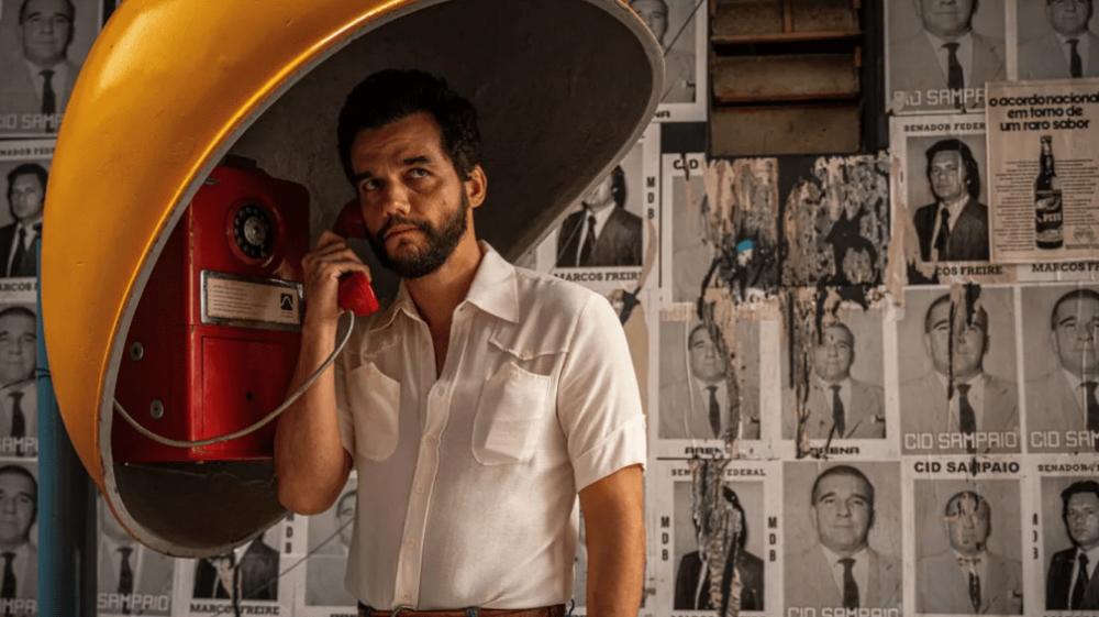 Ator Wagner Moura em um orelhão de rua, segurando o telefone na orelha direita, em uma cena do filme O Agente Secreto.
